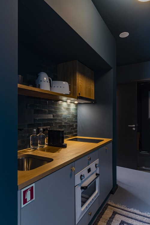 Kitchenette moderna em estúdio de hotel no Porto com balcões de madeira, painel de azulejos escuros, eletrodomésticos em aço inoxidável e cafeteira SMEG