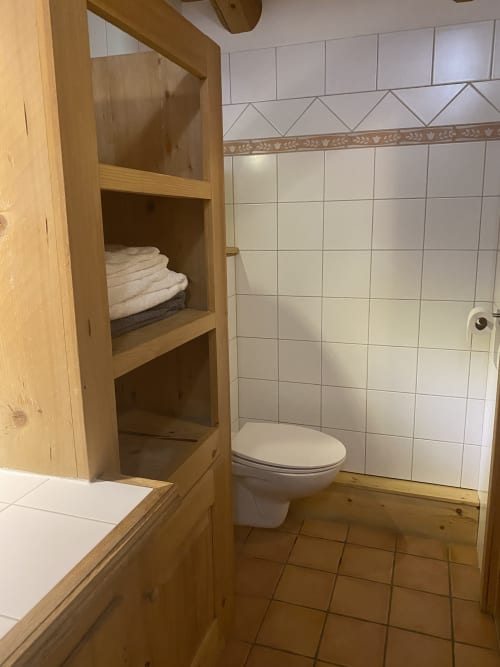 Salle de bain de l'appartement Vorès à La Ferme des Georgières avec toilettes, carrelage blanc, étagères en bois avec serviettes et