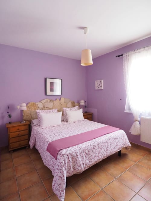 Dormitorio púrpura brillante con ropa de cama floral, mesillas de madera, lámpara colgante y vistas al jardín en zona rural española