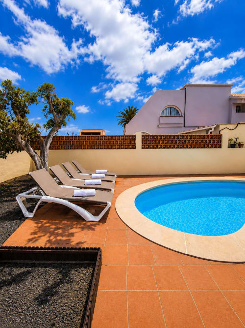 Patio soleado junto a la piscina de la Villa Paloma, Corralejo, con tumbonas y edificio rosa