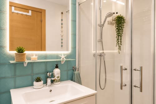 Salle de bains moderne aux carrelages turquoise, miroir éclairant, lavabo blanc et douche vitrée avec plante retombante à Sara