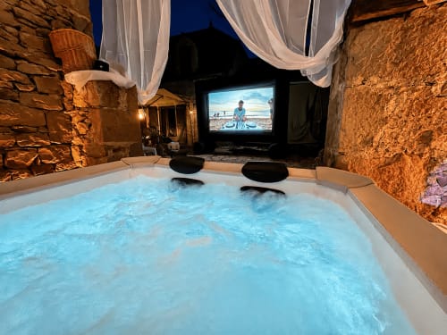 Baignoire spa chauffée aux bulles scintillantes dans une cour en pierre la nuit, écran de cinéma privé affichant une scène de plage, Domaine Valenquercy