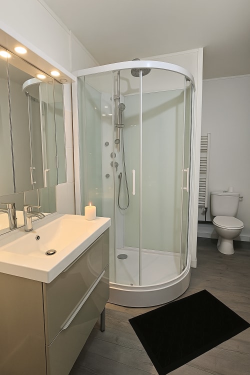 Salle de bain moderne avec douche à l'italienne vitrée incurvée, vasque blanche, miroir et toilettes à l'appartement de Saint-Valery-en-Caux