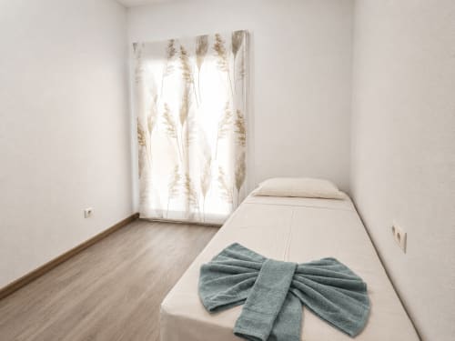 Habitación luminosa en Apartamentos El Calón Playa con cama individual, toalla doblada y ventana con cortinas de estampado botánico