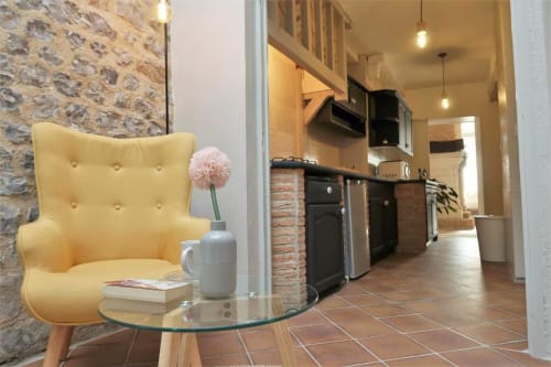Fauteuil jaune avec un vase de fleurs roses à côté d'un mur en pierre, cuisine moderne visible par la porte de l'appartement La Suite