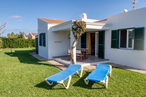 Villa blanca con persianas verdes y tumbonas azules en jardín cuidado de Las Golondrinas, Cap d'Artrutx