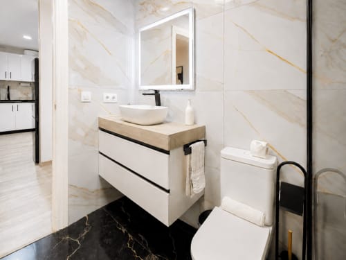 Baño moderno con paredes de mármol, vanidor blanco, lavamanos suspendido, espejo iluminado y piso de mármol negro en Apartamentos El