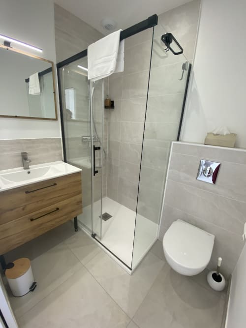 Modern bathroom at La Maison Blanche with glass shower enclosure, white toilet, wooden vanity with sink, and tiled floors