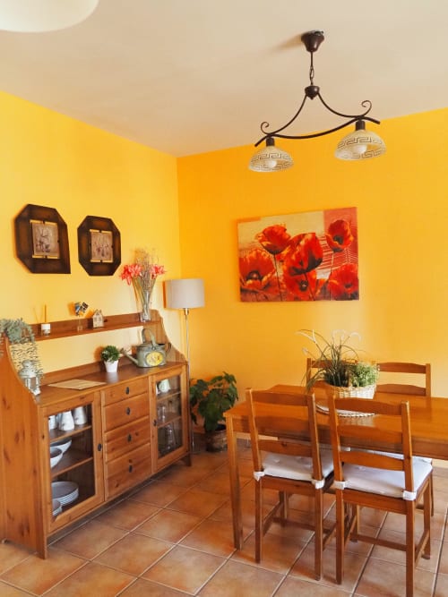 Comedor luminoso en amarillo brillante con muebles de madera, cuadro de amapolas rojo y lámparas colgantes en el apartamento Senda del Duratón