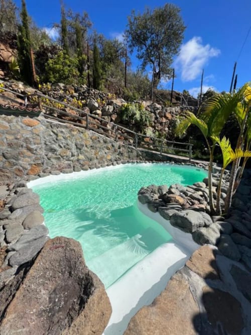 Piscina de borde infinito color turquesa rodeada de rocas volcánicas en Villa Awara, La Palma, con plantas tropicales y vistas a las montañas