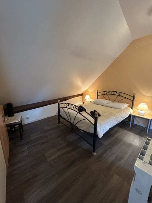Chambre mansardée avec lit en métal, draps blancs, lampes de chevet et parquet foncé à Hébergement
