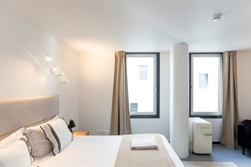 Quarto estúdio moderno em hotel do Porto com cama, janelas, cortinas bege, almofada, frigorífico e luz natural