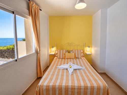 Luminoso dormitorio frente al mar con pared amarilla, ropa de cama a rayas y vistas al mar en Apartamentos El Calón Playa, Cuevas del