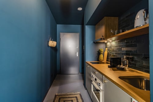 Kitchenete moderna em estúdio de hotel no Porto com paredes azuis, balcões de madeira, backsplash preto e eletrodomésticos em aço inoxidável