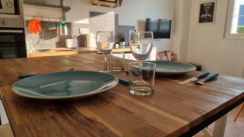 Dining table set for two at Ô LOCH apartment in La Fôret-Fouesnant with teal plates, glassware, and kitchen view