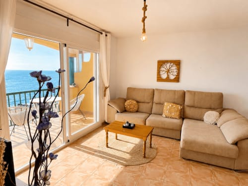 Bright beachfront apartment living room with tan sofa, sea view balcony, wooden table, and coastal decor at Apartamentos El