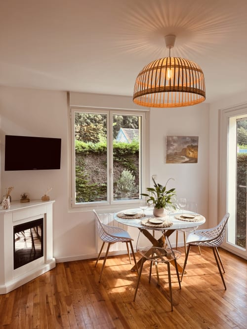 Salle à manger lumineuse du cottage Bobochic à Étretat, table ronde, luminaire suspendu tressé, cheminée et vue sur le jardin