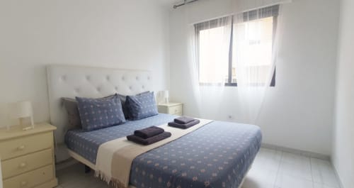 Camera da letto luminosa nell'appartamento Medano Center vicino alla spiaggia El Médano, con biancheria blu decorata, testiera bianca e vista sulla città