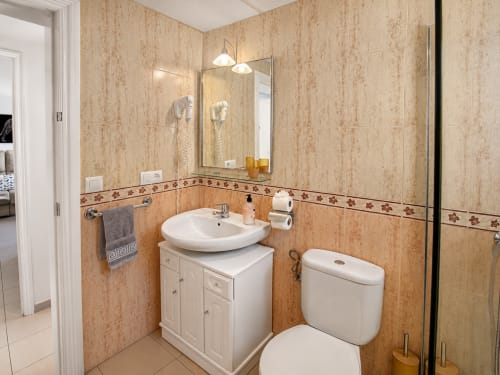 Moderno baño con azulejos beige, lavamanos blanco, inodoro, espejo con lámparas colgantes en Apartamentos El Calón Playa