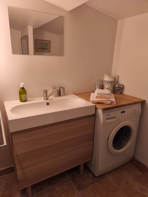 Salle de bain moderne avec meuble vasque en bois, lavabo blanc, machine à laver et miroir à l'appartement Magnanerie de Sainte-Croix.