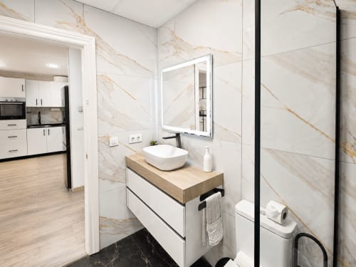 Baño moderno en Apartamentos El Calón Playa con paredes de mármol, lavamanos blanco, espejo iluminado e inodoro
