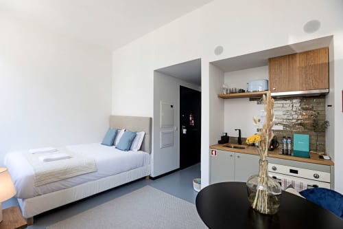 Estúdio compacto no Porto com cama de almofadas azuis, kitchenette com armários de madeira e área de refeições