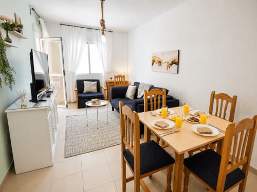 Luminoso salón diáfano con mesa de comedor de madera, sofá negro, televisor y cocina moderna en Apartamentos El