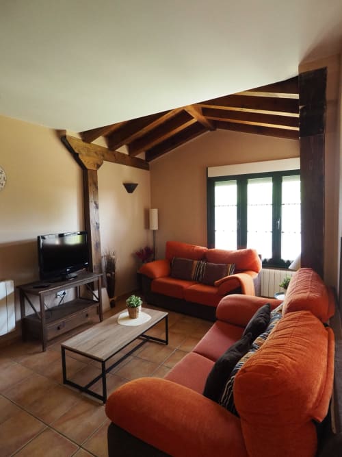 Amplio salón con sofás naranjas, vigas de madera, TV y vistas al jardín en Senda del Duratón
