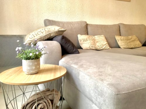 Canapé sectionnel beige avec coussins décoratifs et table d'appoint ronde en bois avec fleurs en pot dans l'appartement Le Charmant Zola