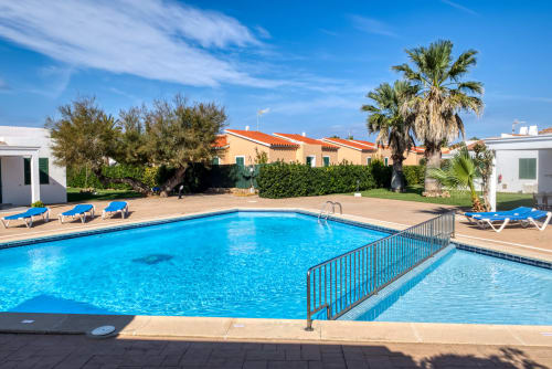Piscina del resort Las Golondrinas con tumbonas, palmeras y edificio de tejado naranja en Cap d'Artrutx