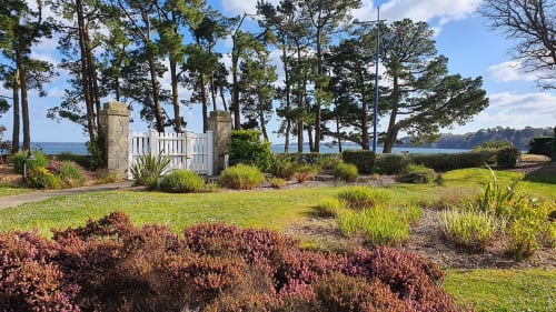 Seaside garden at Cap Coz apartment in La Fôret-Fouesnant with flowering shrubs, manicured lawn, tall pines, and ocean view