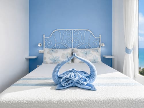 Bright beachfront bedroom with ornate white iron bed, blue accent wall, and decorative blue towel arrangement with sea view