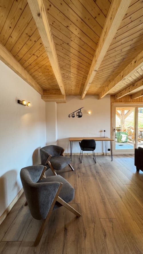 Modern alpine apartment lounge with exposed wooden beams, gray armchairs, desk, and glass doors opening to terrace at La
