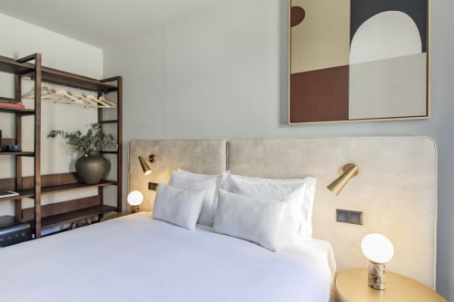 Quarto estúdio moderno e compacto na Benefit Collection Porto com cabeceira estofada bege, roupas de cama brancas e arandelas em latão