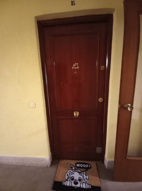 Dark red wooden door entrance to apartment in Benavente with "WOOF" dog doormat on ground floor