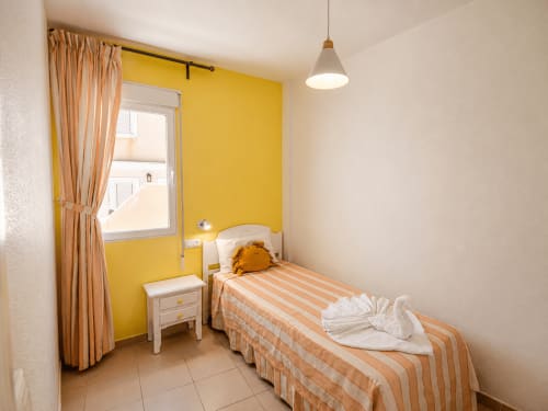 Dormitorio luminoso con pared de acento amarilla, cama individual con ropa de cama a rayas, ventana con cortinas naranjas, luz colgante y