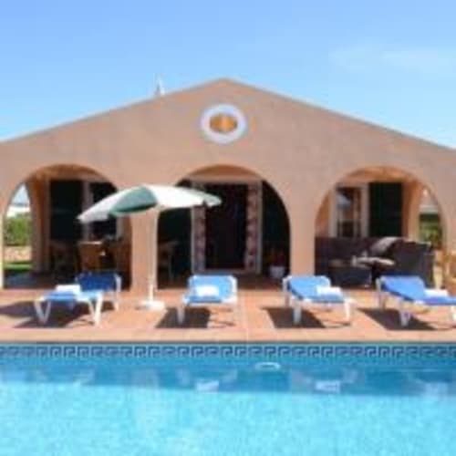 Zona de piscina de villa Las Golondrinas con tumbonas, sombrilla y edificio de terracota en Cap d'Artrutx
