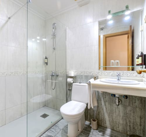 Baño moderno con ducha abierta, azulejos blancos, espejo, lavamanos e inodoro en Hostal El Molino, Collado Villalba