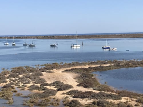 Sailboats anchored in calm blue waters near Tavira with sandy marshland and distant coastline