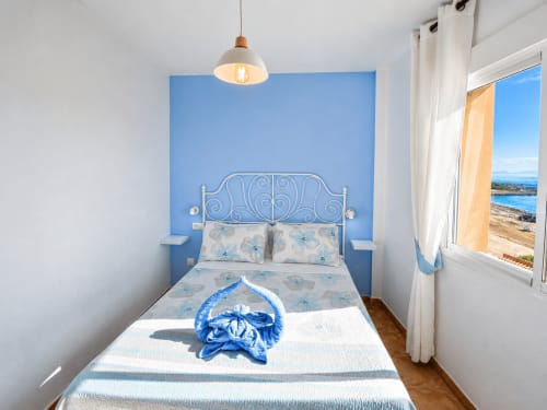 Bright bedroom with blue accent wall, white metal bed frame, sea view window at Apartamentos El Calón Playa, Cuevas del