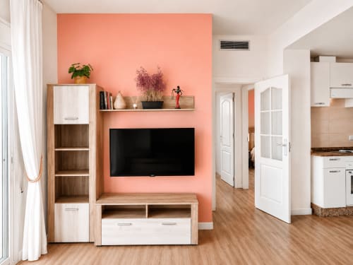 Modern apartment living room with coral accent wall, flat-screen TV, wooden storage unit, and kitchen access at Apartamentos