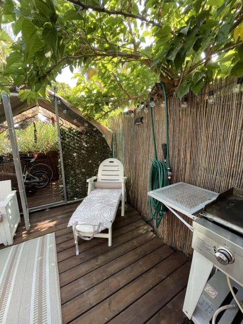 Terrasse de jardin ombragée à La Montagnette Cap d'Agde avec transats, mobilier blanc, barbecue, tuyau enroulé et verdure luxuriante