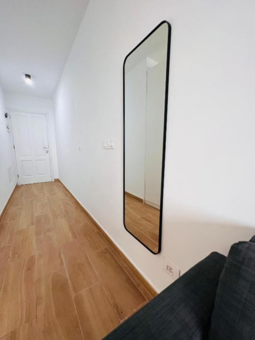 Pasillo moderno con piso de madera, paredes blancas, espejo con marco negro, puerta blanca y vista parcial de sofá oscuro en La Palma