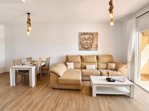 Modern apartment living room with tan sofa, white dining table, wooden floors, and sea view at Apartamentos El Calón Playa