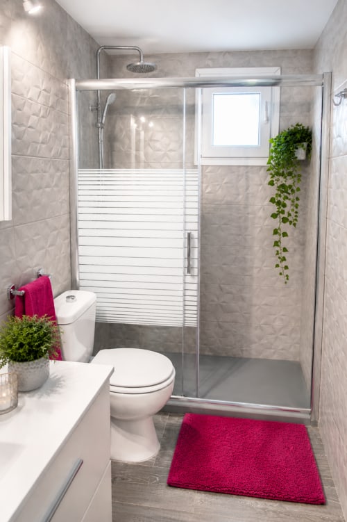 Salle de bain moderne avec carrelage gris, douche vitrée, toilettes blanches, tapis de bain rose et plante verte chez Sara & Gino