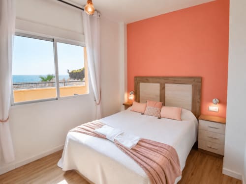 Bright bedroom with coral accent wall, sea view balcony, wooden bed frame, and modern furnishings at Apartamentos El Calón