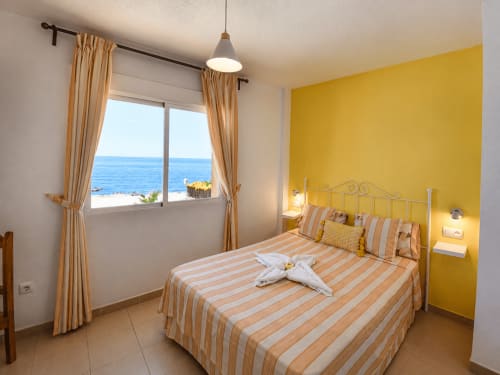 Dormitorio amarillo brillante con ropa de cama rayada, ventana con vista al mar y terraza costera en Apartamentos El Calón Playa