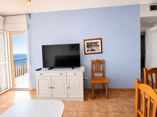 Bright apartment living area with sea view, flat-screen TV on cream cabinet, wooden chairs, blue walls, balcony access