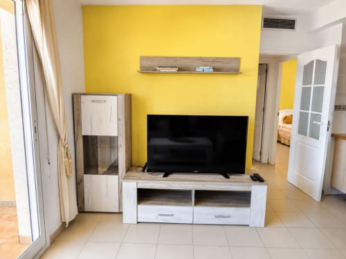 Luminoso salón amarillo con televisor en mueble blanco, estantería de madera y acceso a vistas al mar en Apartamentos El Calón