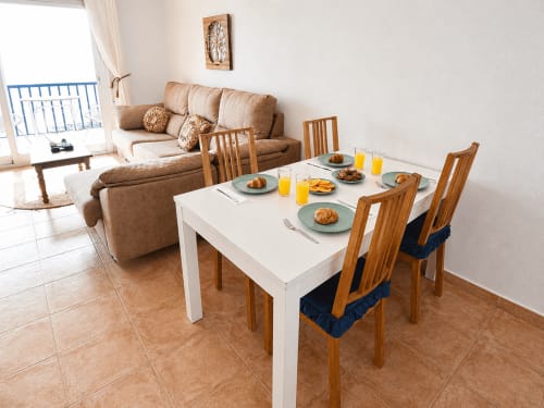 Modern dining area in Apartamentos El Calón Playa with white table, wooden chairs, breakfast setup, and beige sofa